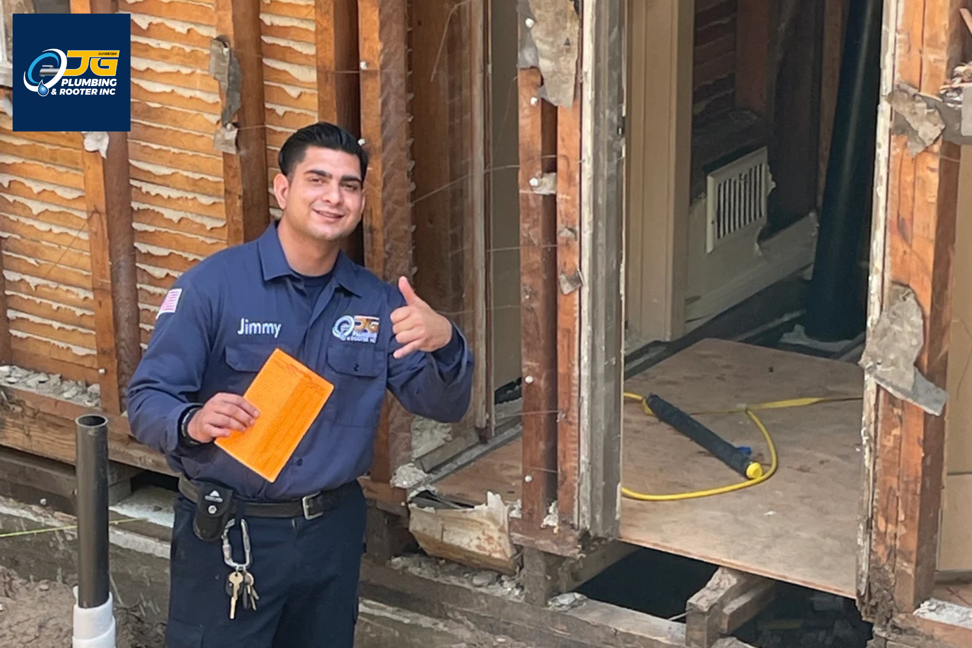 A smiling technician in a blue uniform holds an orange document and gives a shaka sign while standing in front of an exposed wooden wall frame at a construction site.