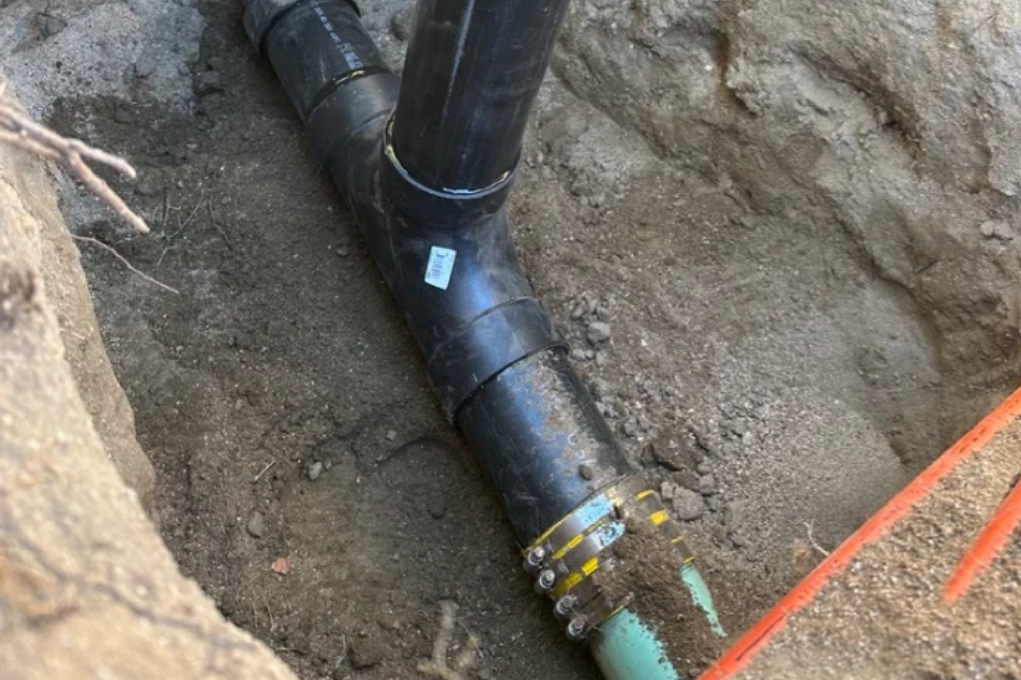 A black ABS plastic T-junction pipe installed in an earthen trench, connected to a green sewer line with a heavy-duty shielded coupling.