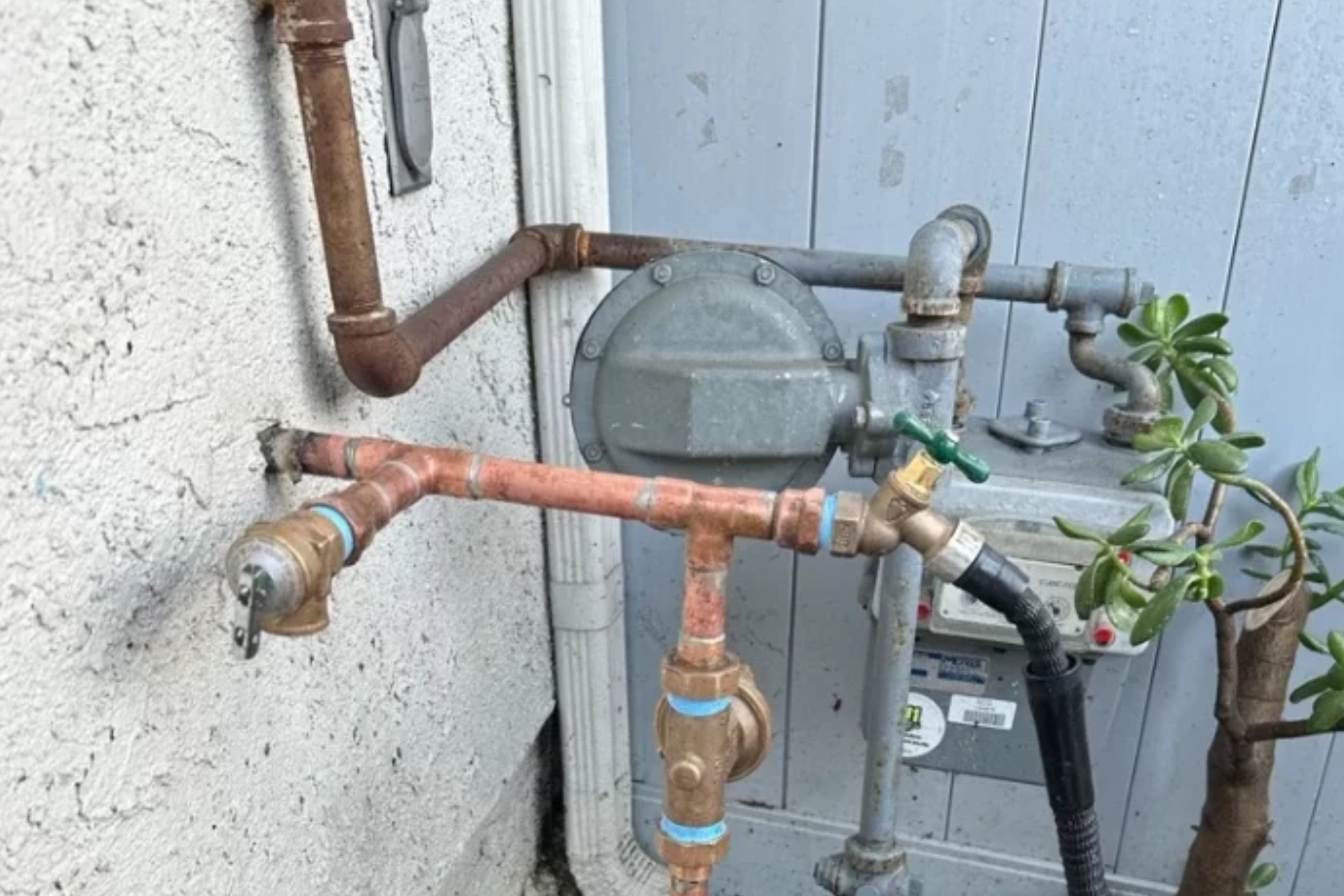 Outdoor residential utility area featuring complex copper and iron plumbing pipes connected to a gas meter and an exterior faucet with an attached hose.