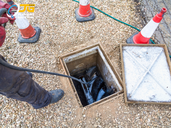 A worker feeds a professional drain snake cable into an open outdoor sewer access point surrounded by gravel and orange traffic cones.